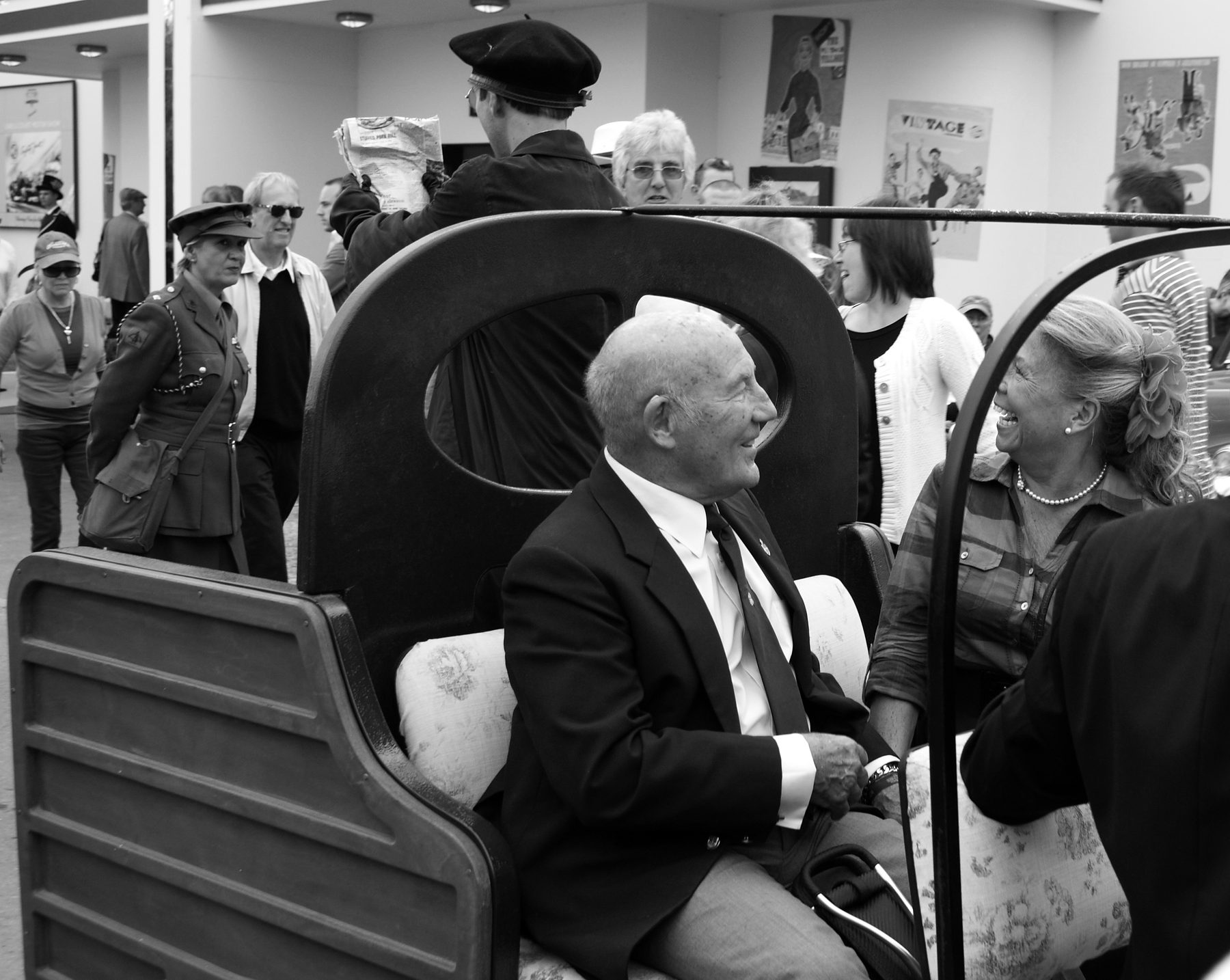 An elderly man in a blazer and tie sits laughing in an open wooden carriage, a smiling woman with a flower in her hair facing him, people in period costume behind.
