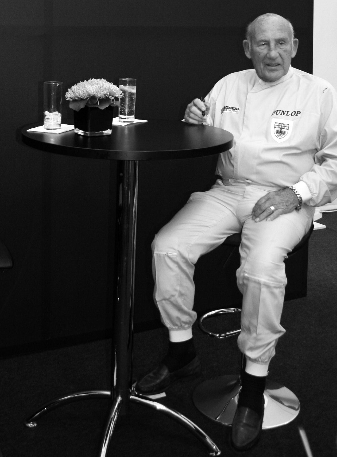 An elderly, balding man in pale racing overalls, the word Dunlop and a club badge with a Union Jack on the chest, sits on a high stool beside a tall round black table with two glasses and a small bowl of flowers, a pen in his right hand.
