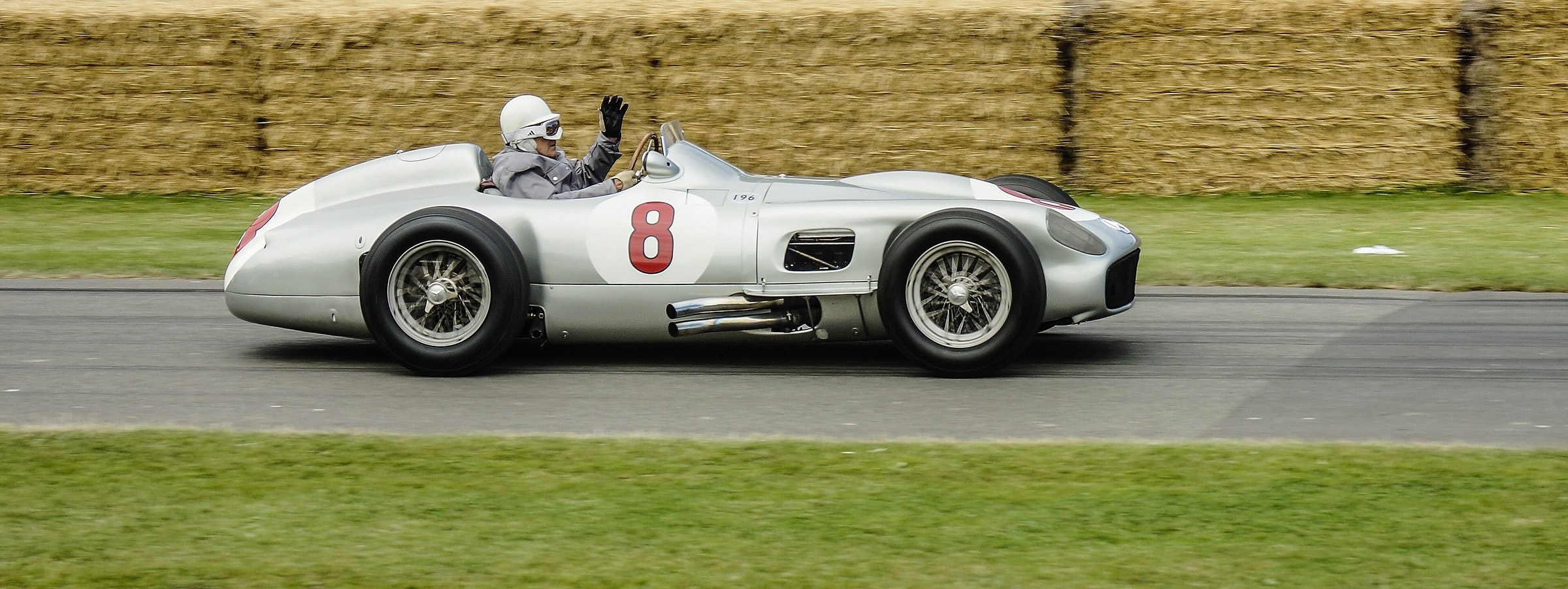 A panned photograph of a silver front-engined racing car numbered 8 running past straw bales, its driver in a white helmet and goggles raising a gloved hand.