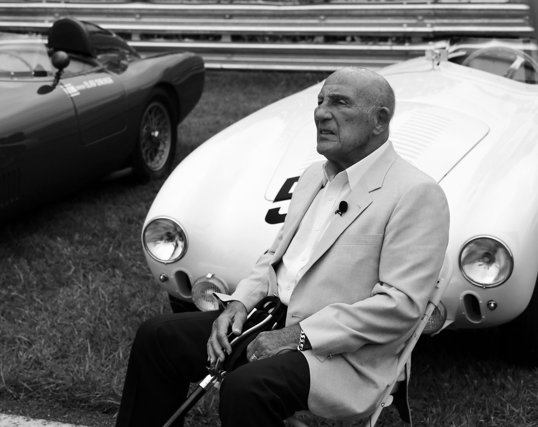 An elderly man in a light jacket sits on a folding chair, a walking stick in his hands, in front of a small open sports car with round headlamps; another sports car is parked on the grass beside it.