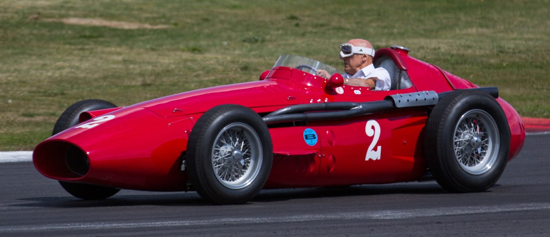 A bare-headed elderly driver in white goggles and a white shirt drives a long red front-engined racing car, numbered 2, along a track beside a grass verge.