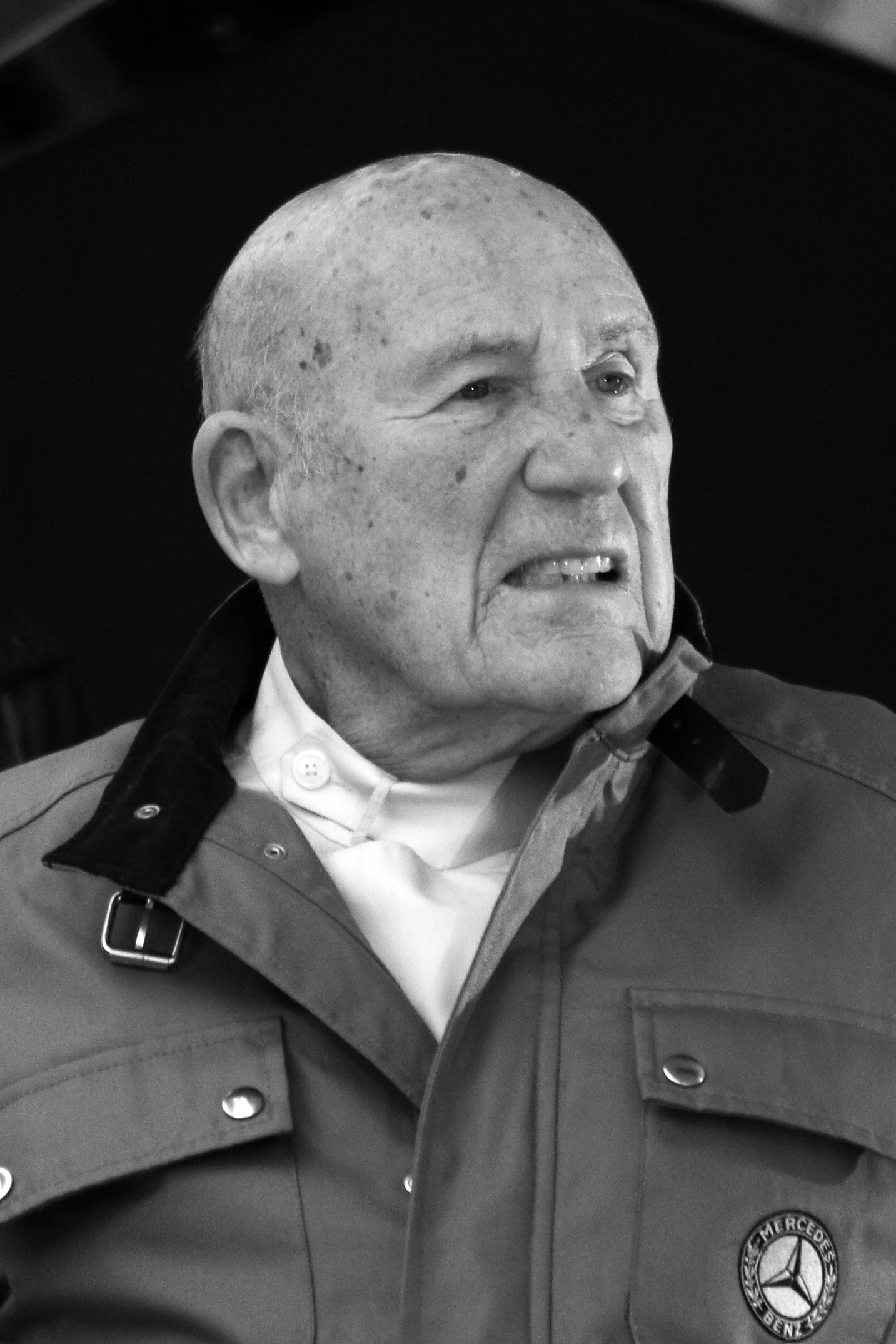 Head and shoulders of an elderly bald man in a dark work jacket with a buckled collar, a Mercedes-Benz badge on its breast pocket, a pale shirt buttoned to the neck; he looks away to the right, his teeth showing.