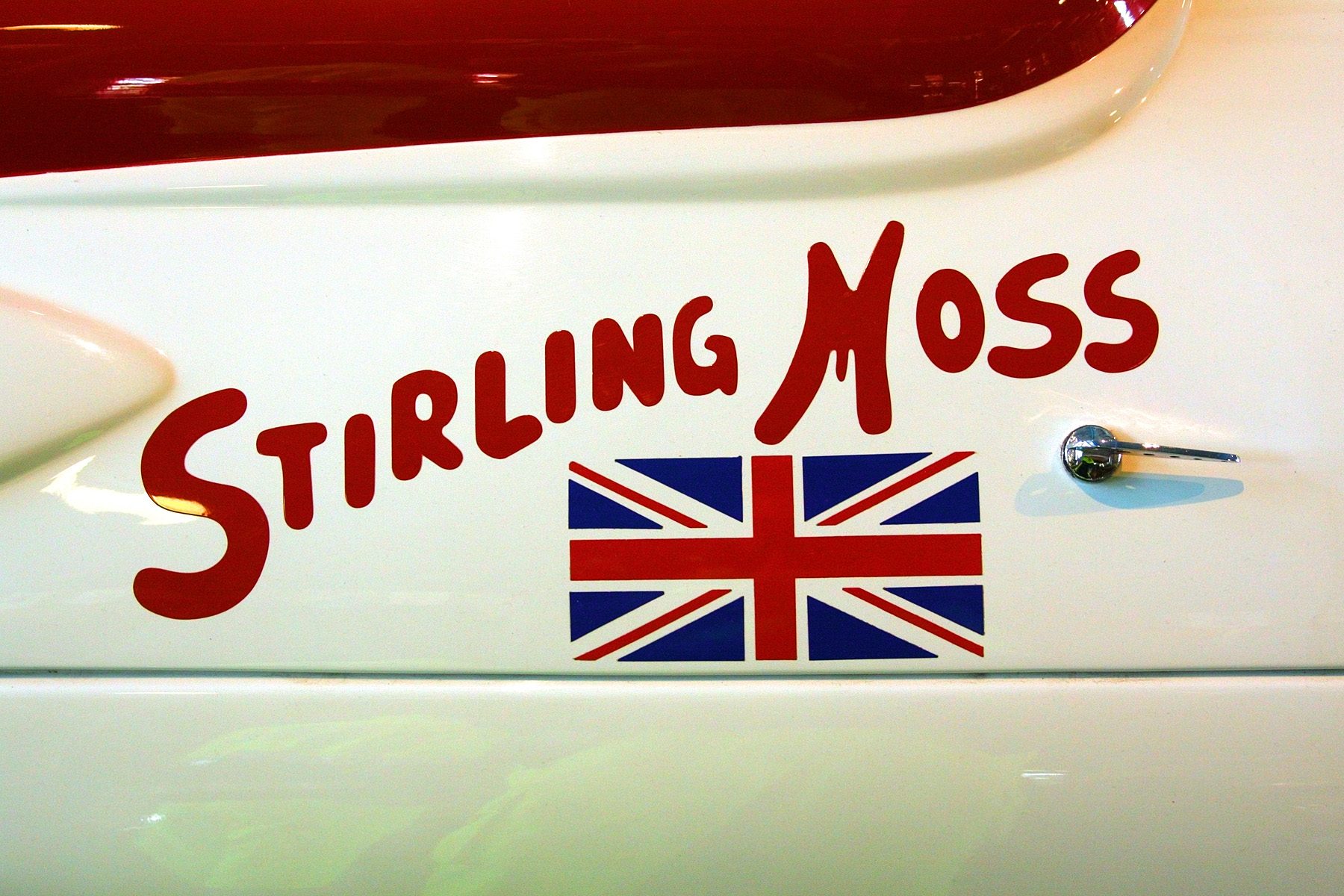 The white flank of a car, lettered ‘Stirling Moss’ in looping red script above a small red, white and blue Union Jack, with a chrome door handle at the right and a band of red bodywork along the top.