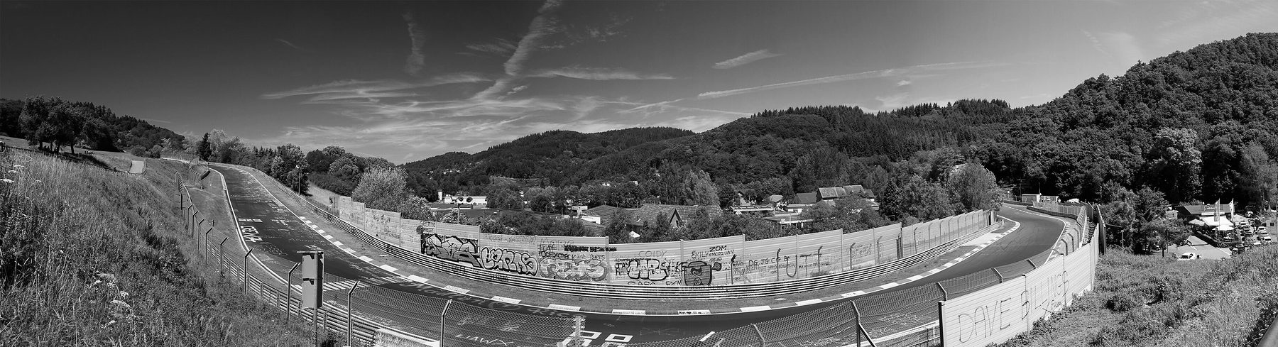 A panorama of a racing circuit sweeping through a valley below wooded hills, past the roofs of a village; catch fences and a painted wall line the track.