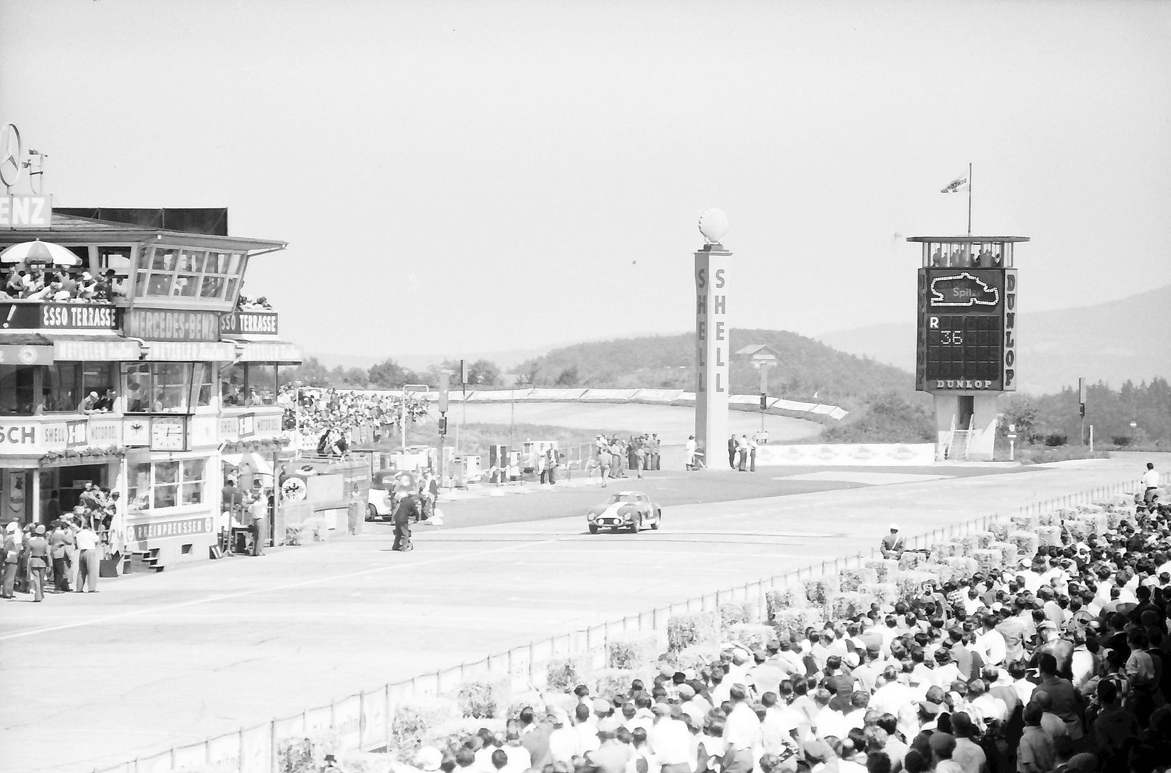 A racing circuit’s straight seen from the stands: a pit building hung with signs at the left, a tall lettered pylon, a lap-board tower with a circuit map, one car on the straight and a deep crowd in the foreground.