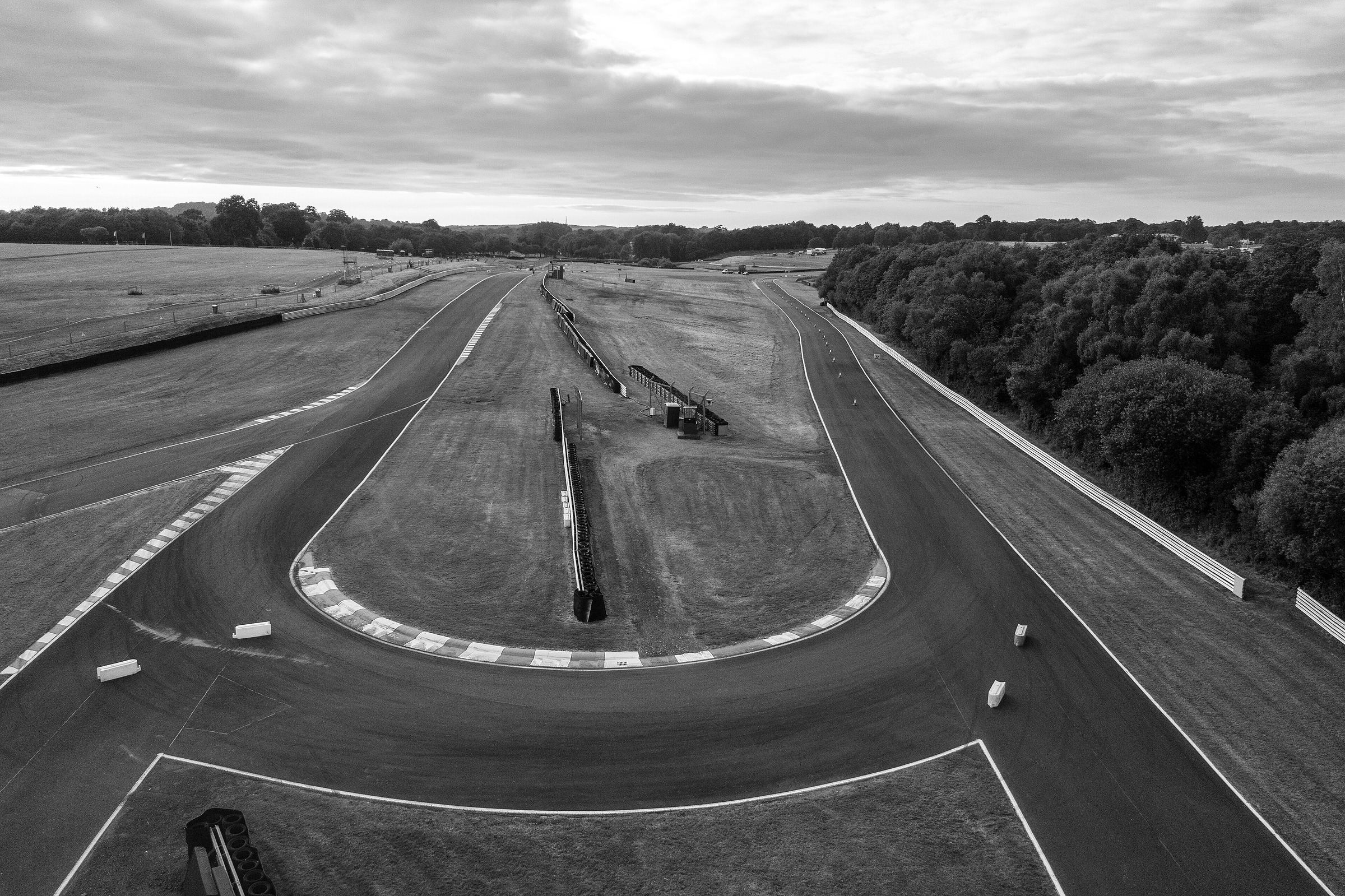 From above, a hairpin bend with striped kerbs, the straights running away from it into wooded countryside under a clouded sky.