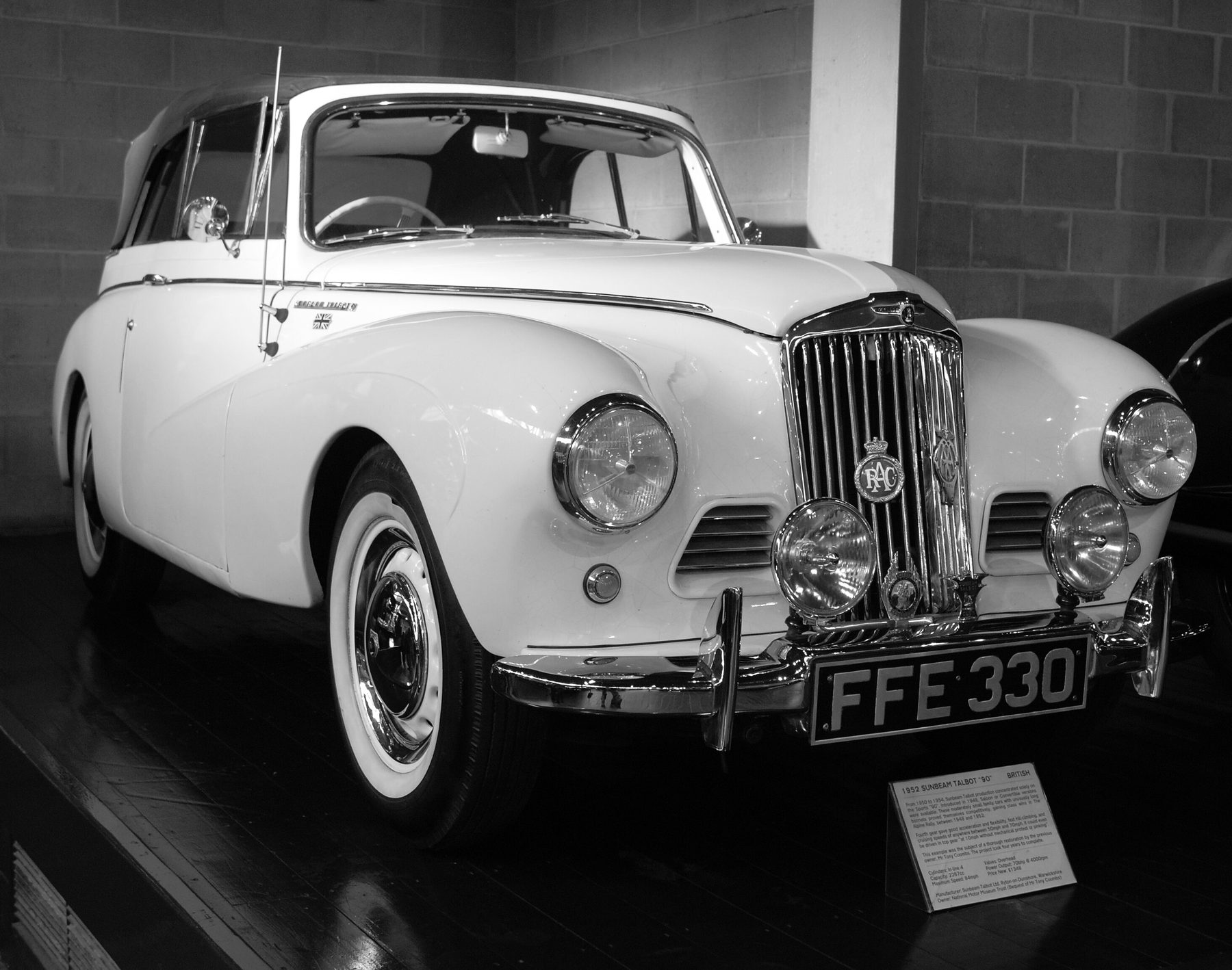 A pale drophead coupé with twin spotlamps and a club badge on its grille stands on a dark plinth in a museum, a brick wall behind it and the edge of a dark car at the right.