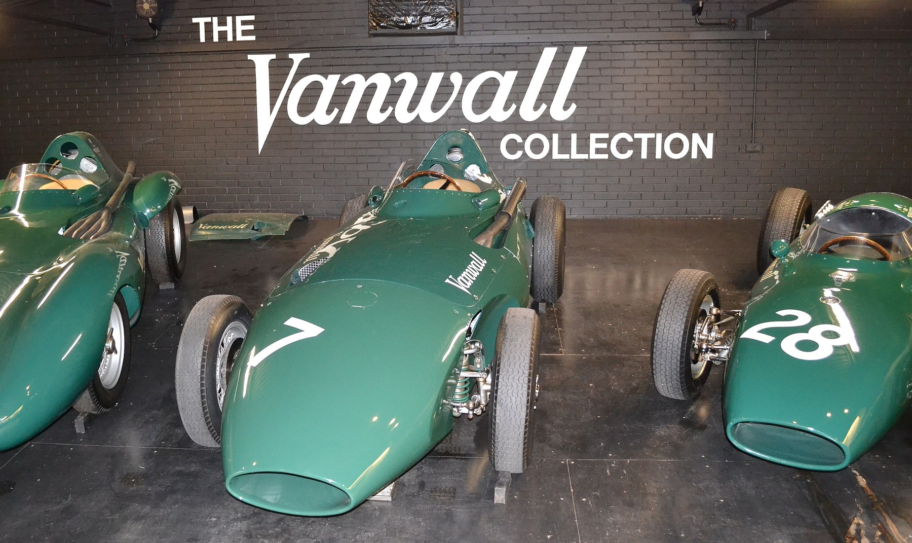 Three green front-engined racing cars stand nose-on in a row on a dark floor under a white wall sign reading ‘The Vanwall Collection’; the middle car is numbered 7, the car at the right 28.