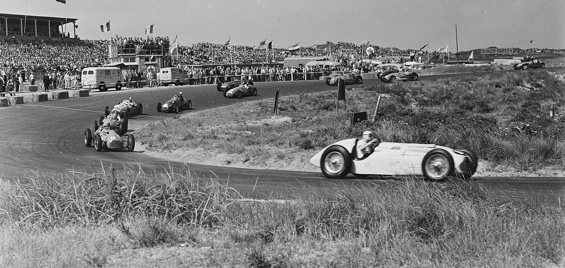 A single-seater with a helmeted driver rounds a bend between banks of dune grass; the rest of the field strings out behind it, past a packed grandstand hung with flags.